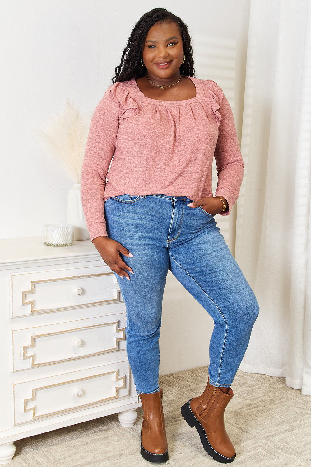 A woman wearing a Double Take Square Neck Ruffle Shoulder Long Sleeve T-Shirt in pink and blue jeans stands with one hand in her pocket, smiling. She has long curly hair and wears a necklace and bracelet. She stands in a room with white decor.