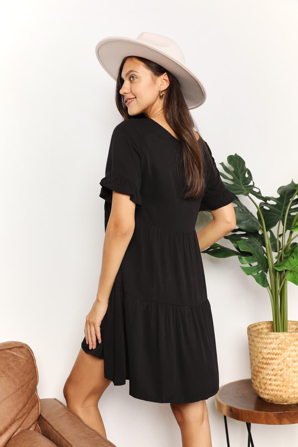 A woman stands indoors wearing the Mandy V-Neck Flounce Sleeve Tiered Dress in navy blue. She has one hand on her hip and is smiling.