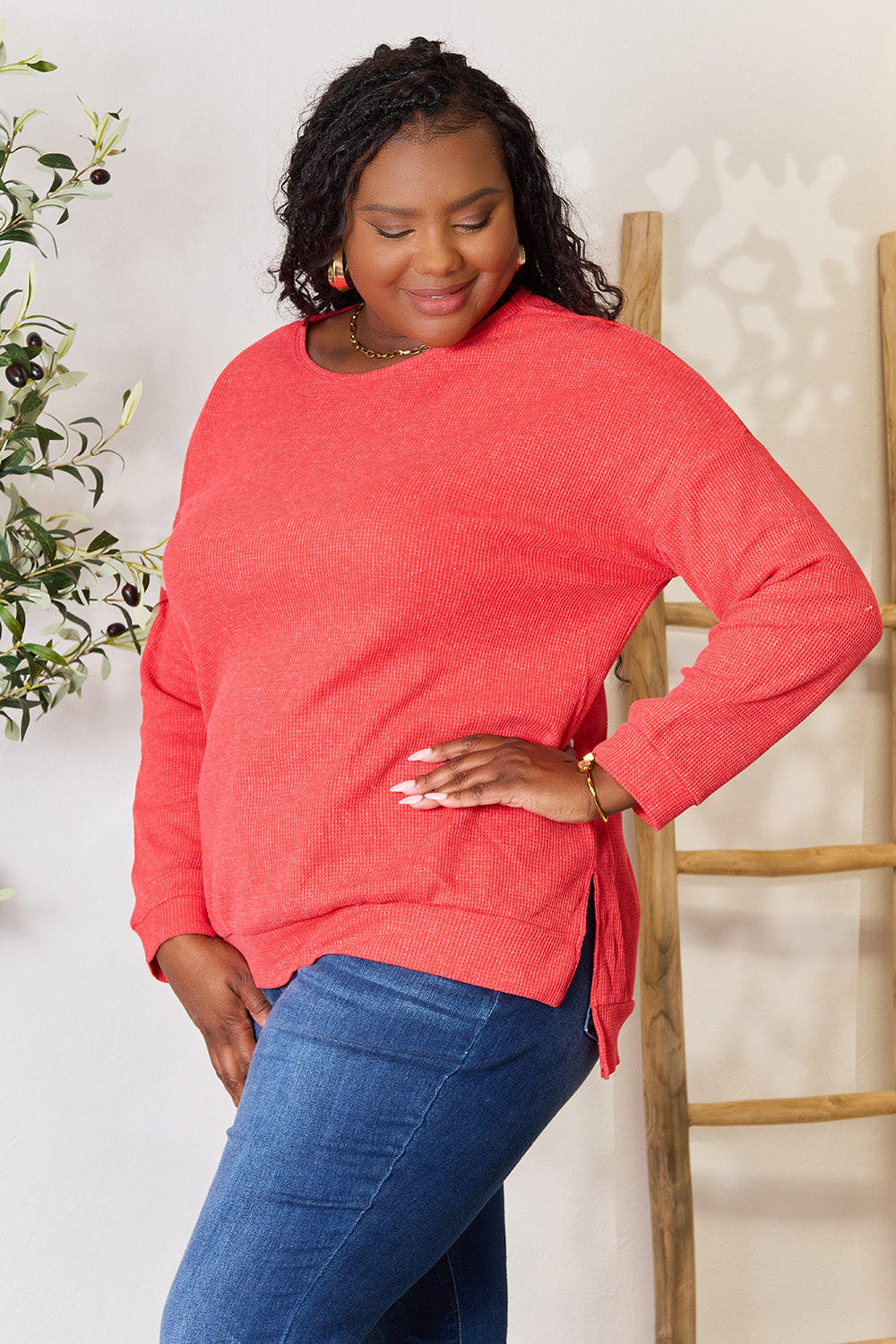 A woman wearing the Basic Bae Round Neck Drop Shoulder Slit Sweatshirt in red and blue jeans stands indoors, smiling and touching her hair, with a decorative plant and wooden ladder in the background.