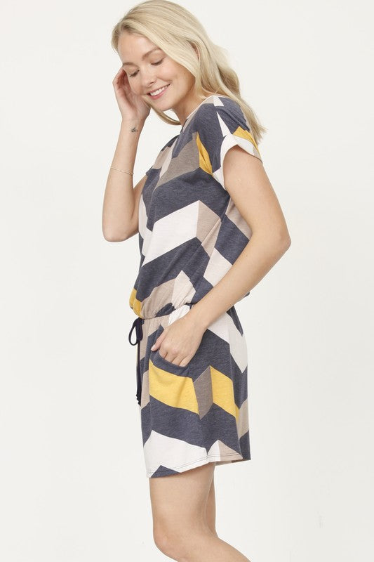 A person is modeling the Plus Drawstring Chevron Romper, featuring a short-sleeve design with a chevron pattern in shades of blue, gray, and beige, set against a plain light-colored background.