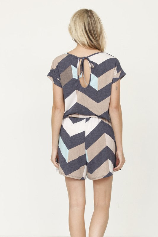 A person is modeling the Plus Drawstring Chevron Romper, featuring a short-sleeve design with a chevron pattern in shades of blue, gray, and beige, set against a plain light-colored background.