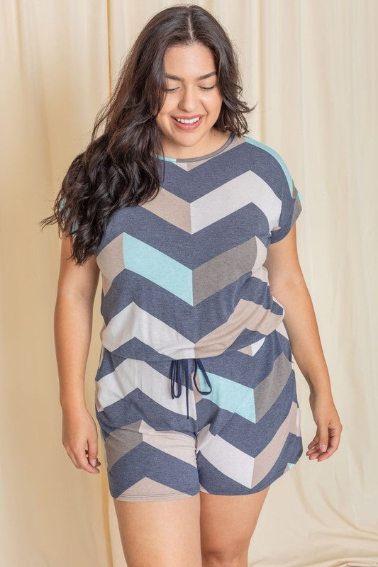 A person is modeling the Plus Drawstring Chevron Romper, featuring a short-sleeve design with a chevron pattern in shades of blue, gray, and beige, set against a plain light-colored background.