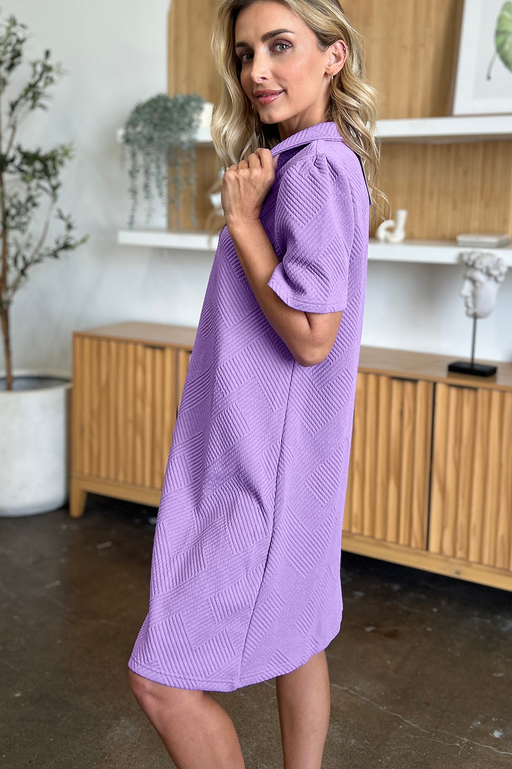 A woman wearing the Double Take Full Size Texture Collared Neck Short Sleeve Dress, featuring a geometric pattern in purple and made from soft polyester, stands against a neutral background. She has long, light hair and is looking slightly to the side. The dress is available in sizes S-3XL and is easy to care for—just machine wash cold.