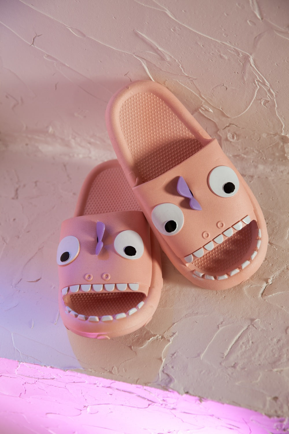 The NOOK JOI Monster Pillow Cloud Slides Non-Slip Slipper features cartoonish monster faces with large eyes, a purple nose, and an open mouth with teeth. Made of durable EVA material, these fun pink cloud slippers also boast an anti-slip design for added safety.