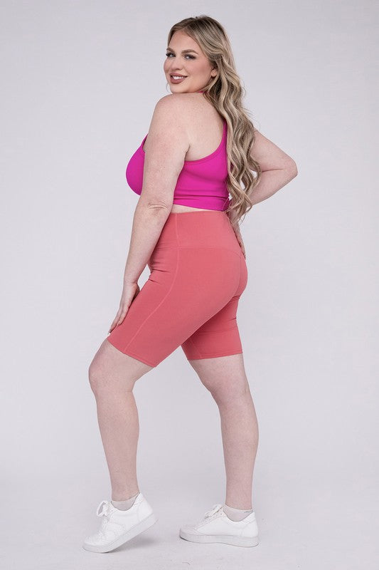 Against a plain background, a person showcases the Plus Athletic High Rise Biker Shorts in coral, crafted from high-quality athletic fabric and paired with a pink top.