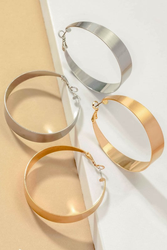 Four Satin Surface Wide Hoop Earrings are displayed on a beige and satin background. Two earrings feature a silver finish, while the other two are gold-plated. Each pair is arranged parallel to one another.