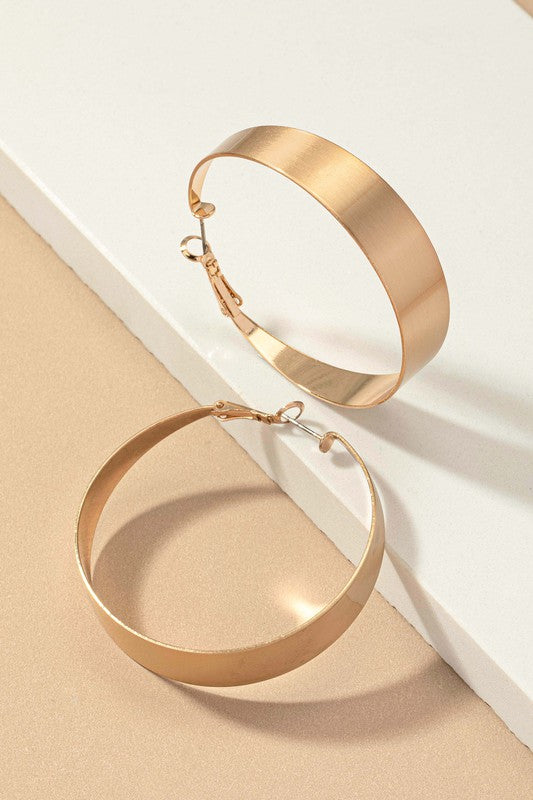 Four Satin Surface Wide Hoop Earrings are displayed on a beige and satin background. Two earrings feature a silver finish, while the other two are gold-plated. Each pair is arranged parallel to one another.