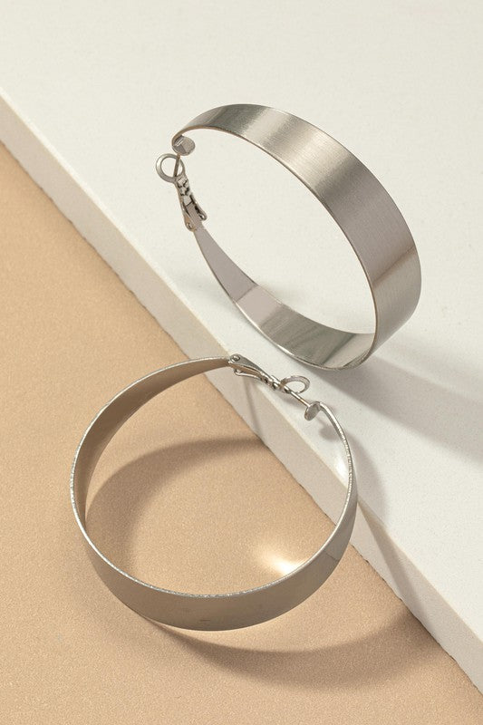 Four Satin Surface Wide Hoop Earrings are displayed on a beige and satin background. Two earrings feature a silver finish, while the other two are gold-plated. Each pair is arranged parallel to one another.