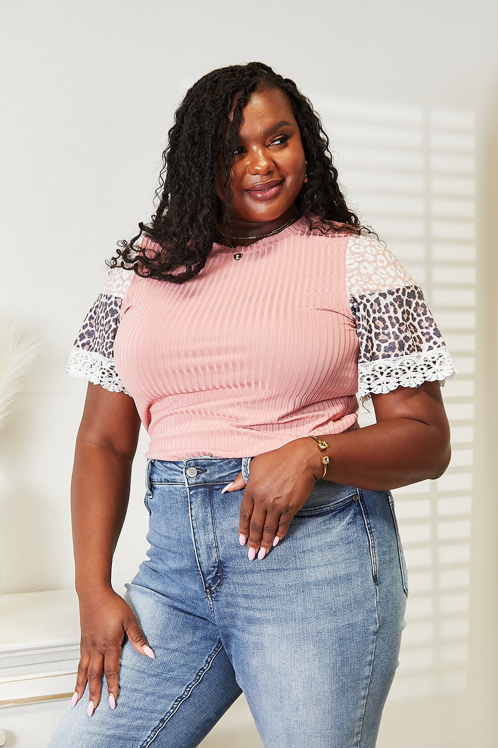 A woman with long hair wearing a trendy Double Take Leopard Lace Trim Ribbed Round Neck Top paired with blue jeans stands indoors smiling.