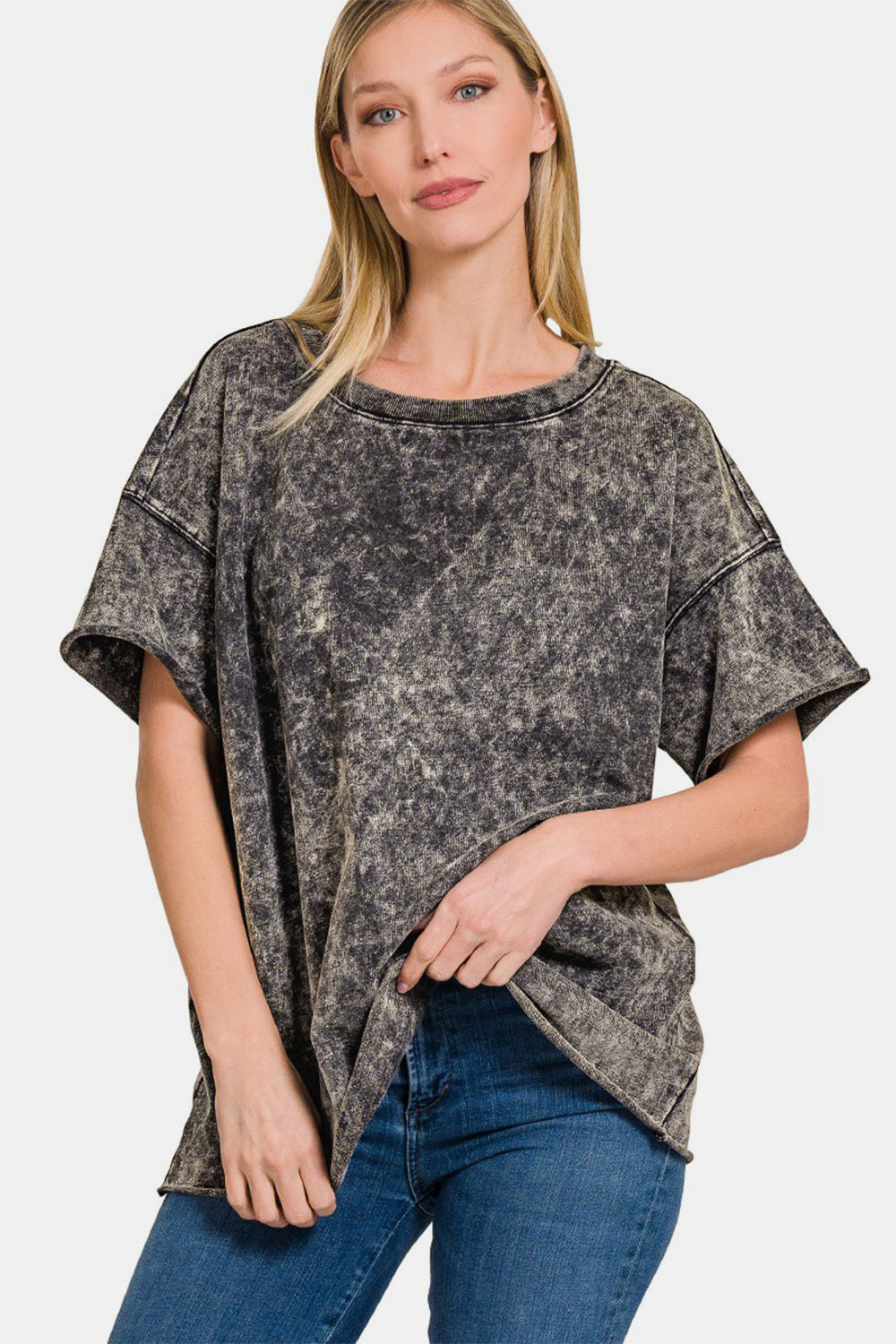 A woman with long blonde hair wearing a Zenana Rolled Round Neck Short Sleeve T-Shirt in grey acid-washed 100% cotton and blue jeans stands against a plain white background.