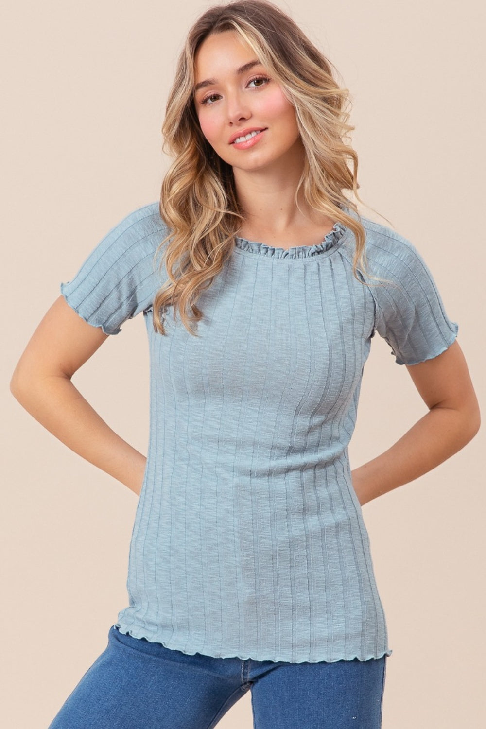 A person wearing the BiBi Ruffle Round Neck Merrow Edge Rib Top, featuring short sleeves and a light blue hue, stands against a neutral background. They have long, wavy hair, with tattoos adorning their arm.