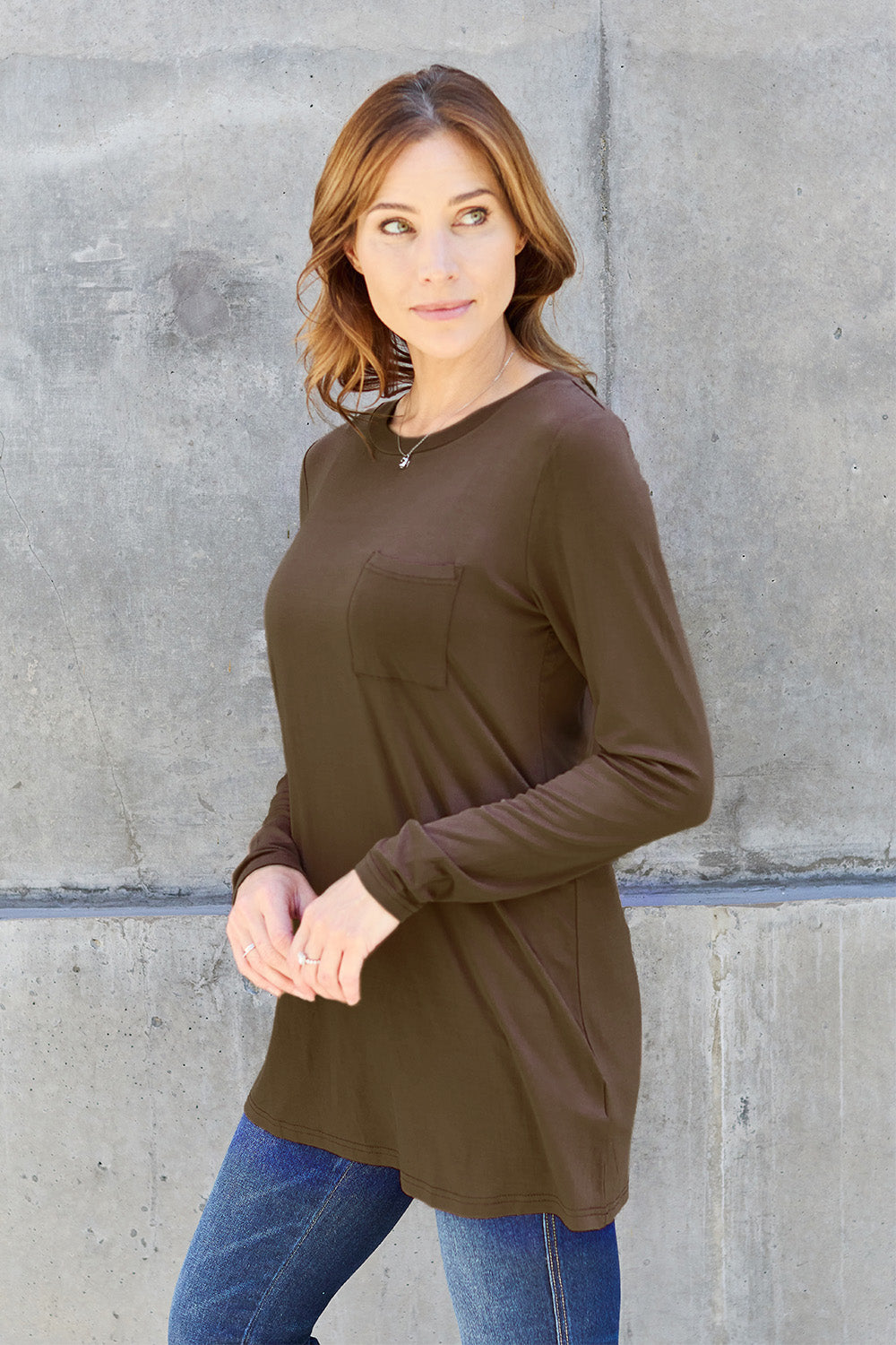 A woman with long hair wearing sunglasses, a Basic Bae Full Size Round Neck Long Sleeve Top in turquoise, and jeans is standing in front of a concrete wall with one hand on her hip.