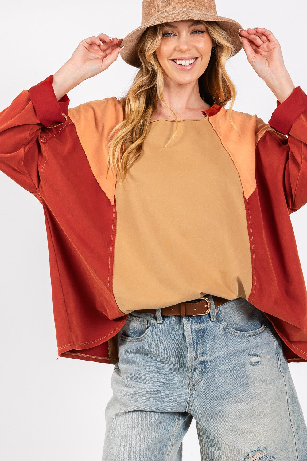 A person in a wide-brimmed hat, wearing the SAGE + FIG Mineral Wash Color Block Top and distressed denim shorts, smiles while adjusting the hat with one hand against a neutral background.