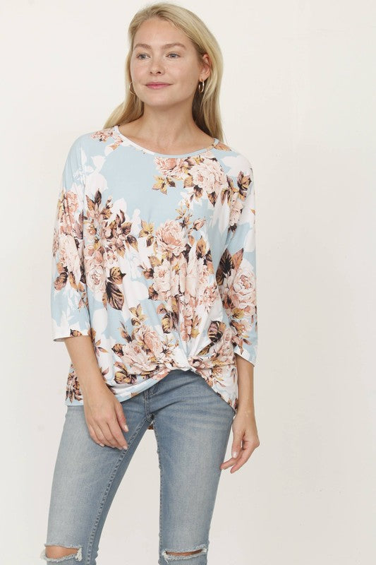 A woman with blonde hair is wearing a Plus Floral Bottom Knot Tunic and blue jeans. Her eyes are closed, and she is touching her hair with her right hand.