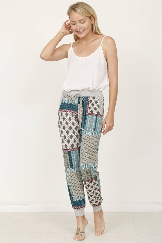 A person is standing indoors, with only part of their upper body visible, wearing a white top and Plus Quilted Print Joggers featuring a colorful pattern. The joggers have a drawstring waist and pockets where the person's hands are tucked. Their left foot is bare.