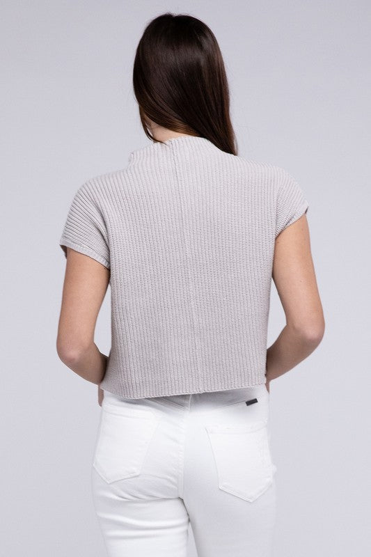 A woman in a JustFab mock neck short sleeve cropped sweater and white pants, smiling slightly, stands with one hand on her hip against a light grey background.