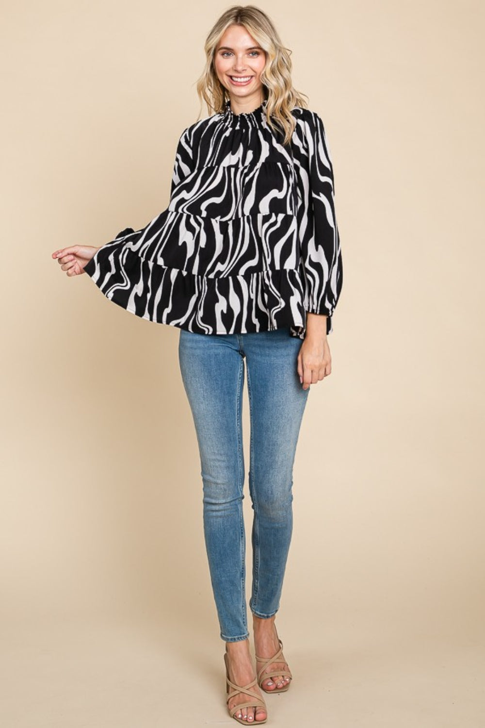 A person wearing a Culture Code Printed Smock Neck Tiered Blouse, featuring a black and white patterned design and long sleeves, paired with blue jeans, stands against a beige background.