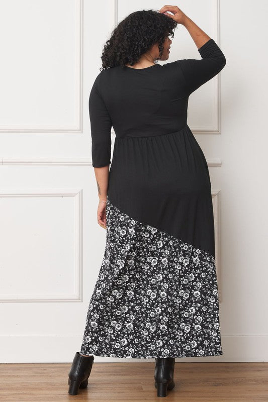 A woman with curly hair is wearing the Asymmetrical Leopard Accent Maxi Dress, featuring a long black design with a floral patterned section at the bottom. She stands against a white paneled wall, her hands tucked in her dress pockets, showcasing its elegant design.