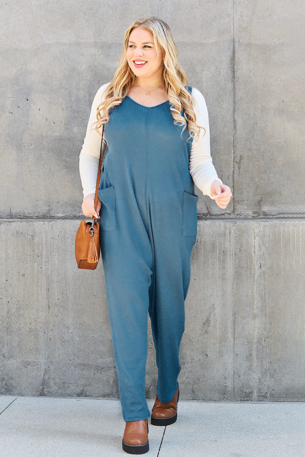 A person stands outdoors wearing the Double Take Full Size Sleeveless Straight Jumpsuit in blue and brown sandals, holding a cream-colored bag. In the background, there's a wooden bench and palm trees.