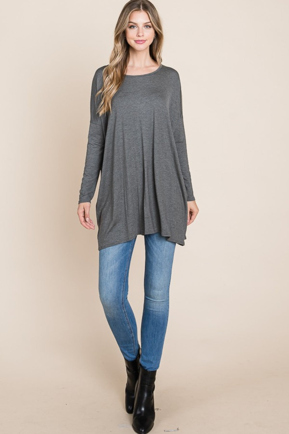 A woman with long hair is wearing a cozy, BOMBOM Round Neck Long Sleeve Oversized Top and blue jeans. She is smiling with one hand touching her head and the other in her pocket. Her comfortable chic wardrobe includes a long necklace, adding a stylish touch to her relaxed outfit.