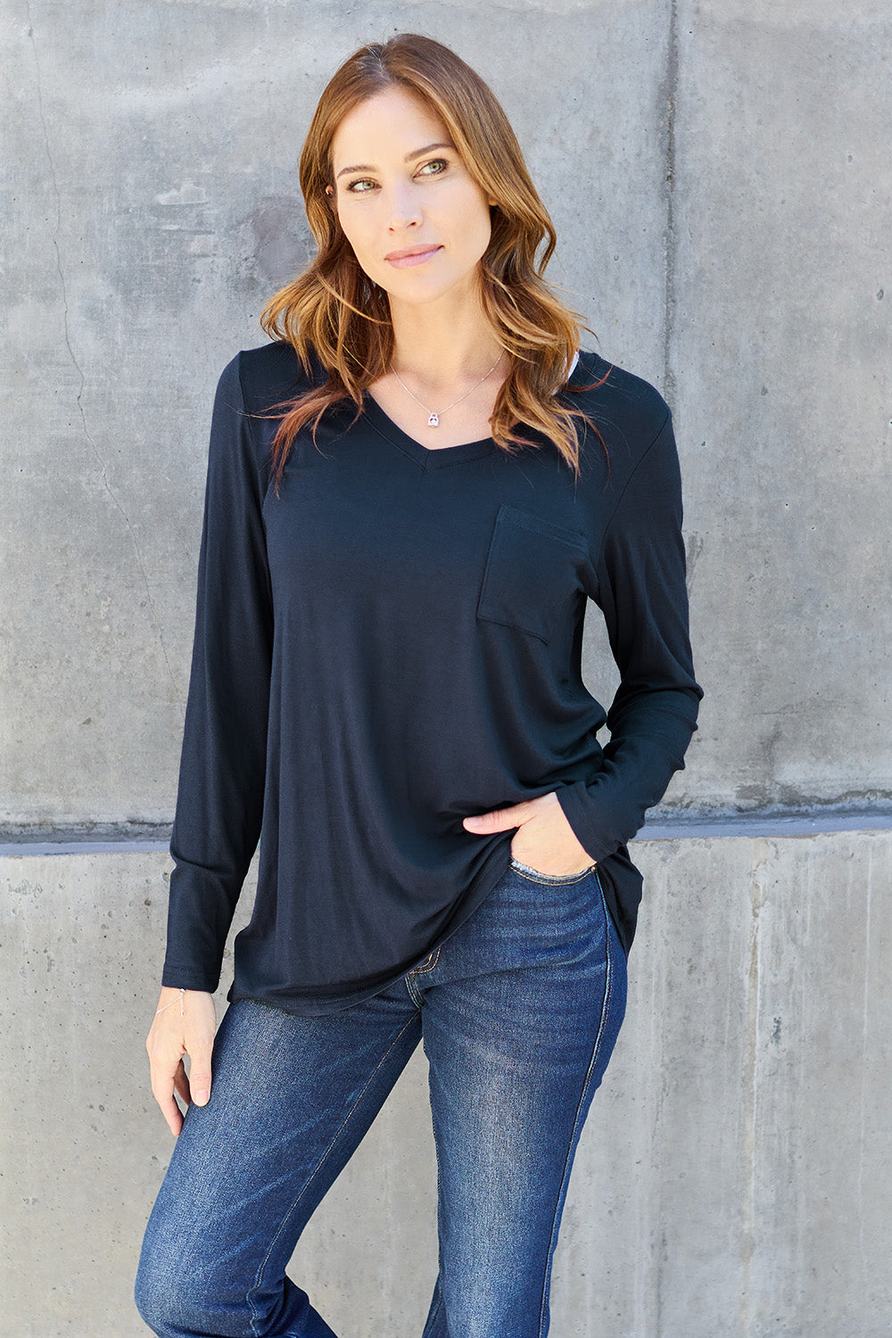 A woman with light brown hair stands against a concrete wall, wearing the Basic Bae Full Size V-Neck Long Sleeve Top in olive green and blue jeans, with a white backpack on her shoulder. Her outfit exudes basic style and is made from polyester, ensuring it’s both comfortable and suitable for machine wash cold. She is looking at the camera.