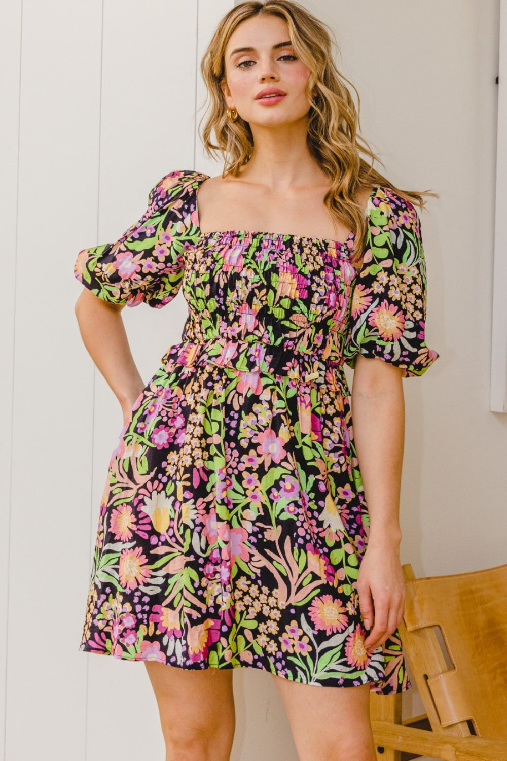 A person stands wearing the ODDI Full Size Floral Tie-Back Mini Dress, a feminine and flirty black floral design with puffy sleeves, perfect for a summer wedding.