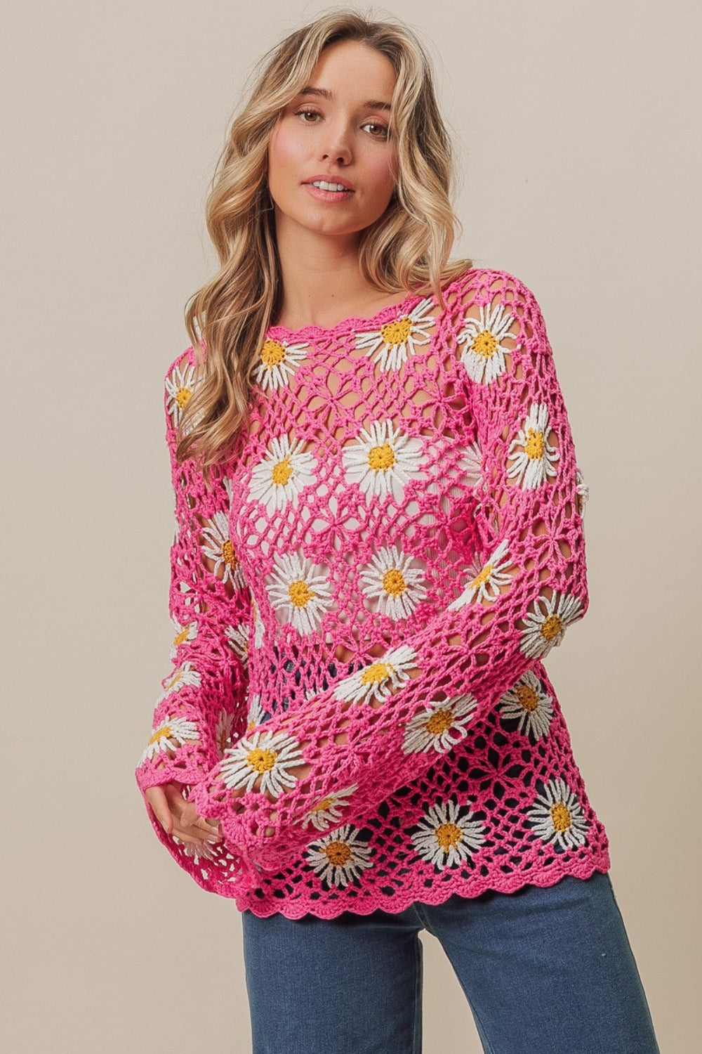 A person stands wearing the BiBi Floral Crochet Net Lace Cover Up, featuring pink crochet work adorned with white and yellow daisy patterns, exuding Bohemian style. They have long, wavy hair and pose with hands together.