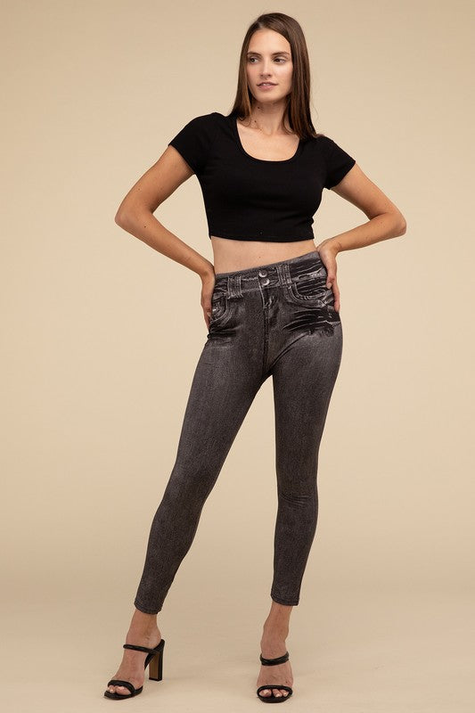 A person wearing Denim Leggings in a high-waisted black design and paired with black heels poses against a plain beige background, highlighting the sleek, stretchy fit.