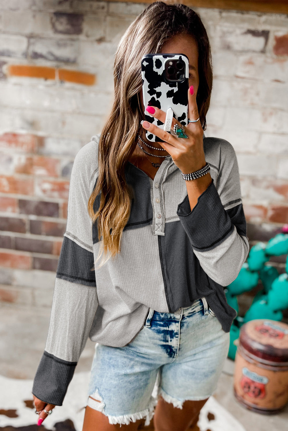 Rear view of a person with long hair wearing a stylish Gray Colorblock Seamed Ribbed Henley Hoodie and light-washed jeans, standing against a brick wall.