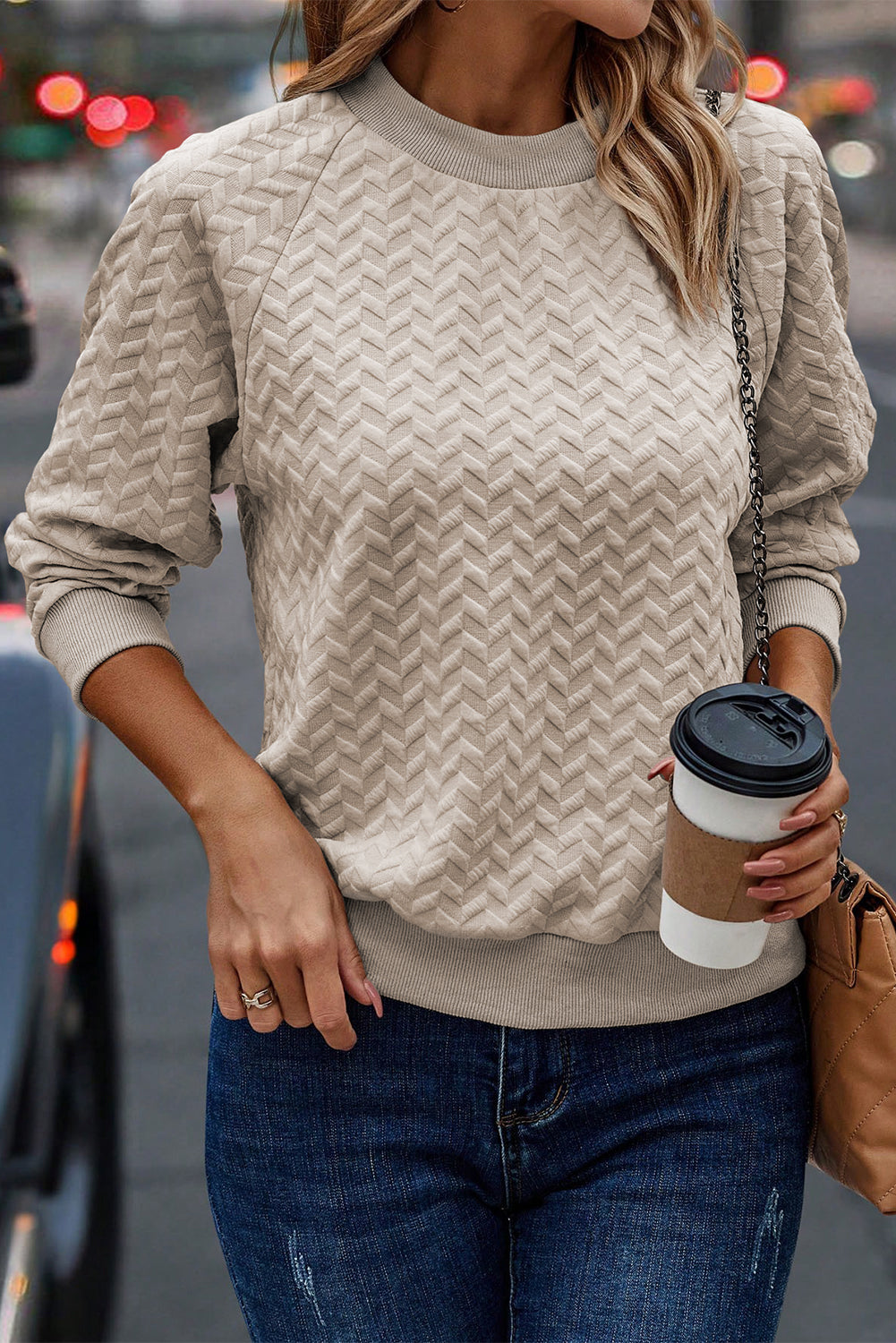 A woman in a Beige Solid Textured Raglan Sleeve Pullover Sweatshirt and blue jeans is looking to the side. She sports a brown hat and earrings, exuding a casual and comfy vibe. Standing next to dried pampas grass against a light background, she seamlessly blends with her textured sweatshirt.