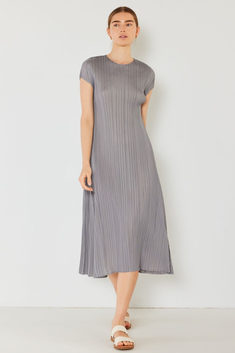 A person stands against a light wall, wearing an elegant Marina West Swim Pleated Cap Sleeve A-Line Dress in black and white sandals.