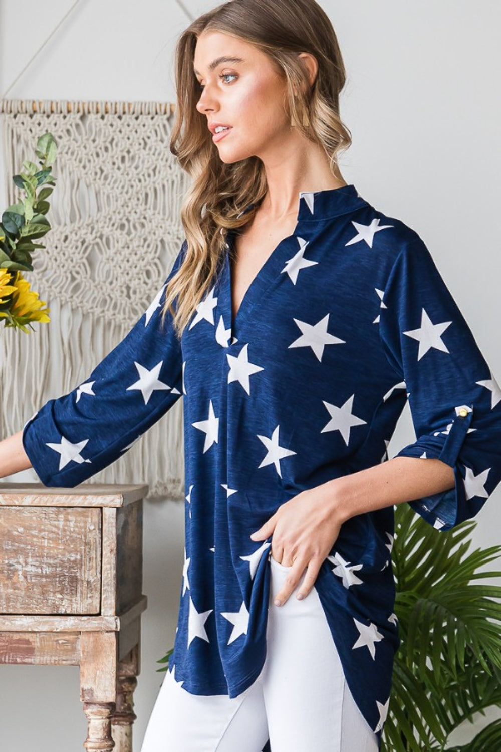 A woman with long hair is sporting the Heimish Full Size Roll-Tab Sleeve Star Print Top in navy blue, paired with white pants, perfect for casual outings. She stands in front of a light-colored wall adorned with round hangings.