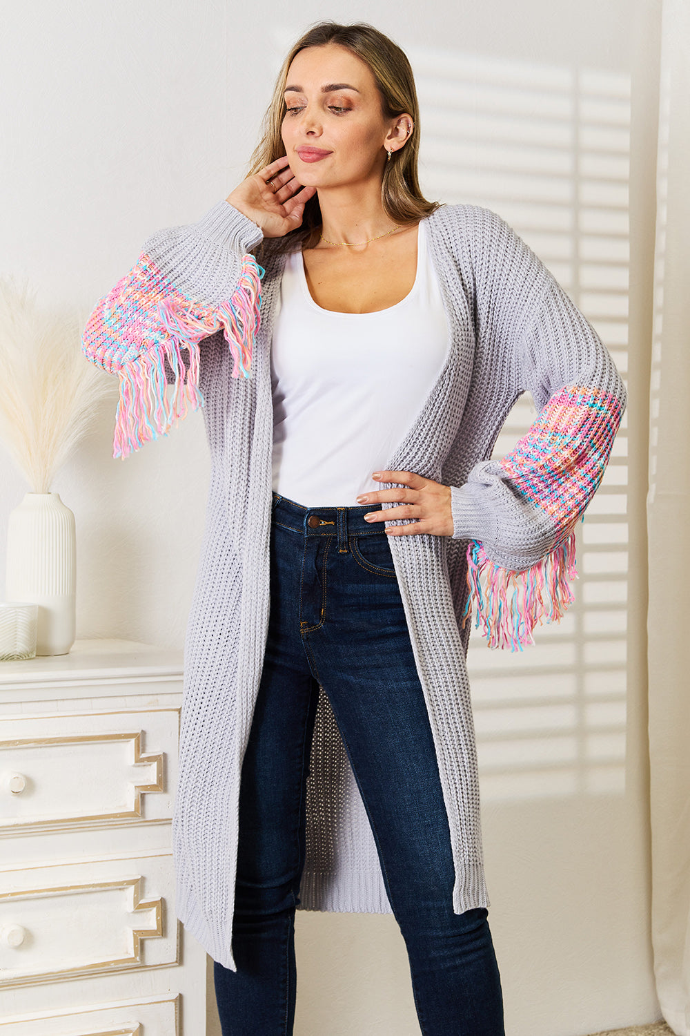 A woman poses indoors wearing the Woven Right Fringe Sleeve Dropped Shoulder Cardigan, a cozy, light gray piece featuring colorful tasselled sleeves. She pairs it with a white top and dark jeans.