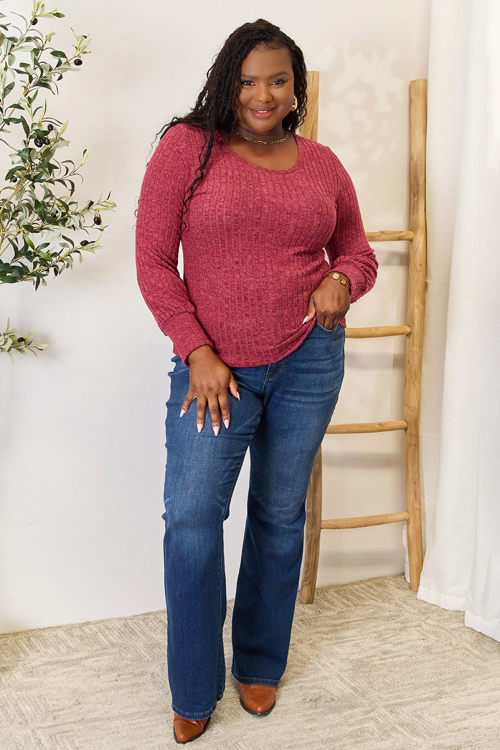 A person in a stylish maroon Ribbed Round Neck Lantern Sleeve Blouse and jeans stands by a wooden ladder, with olive branches in the background.