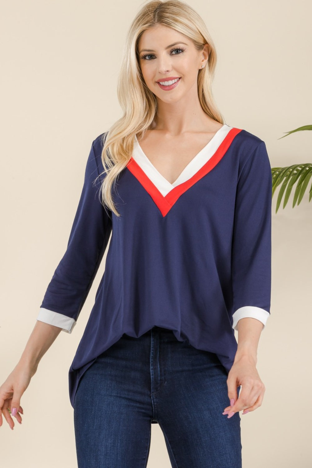 A woman with long blonde hair wearing the Celeste Full Size V-Neck Contrast T-Shirt crafted from lightweight material and dark blue jeans stands smiling.