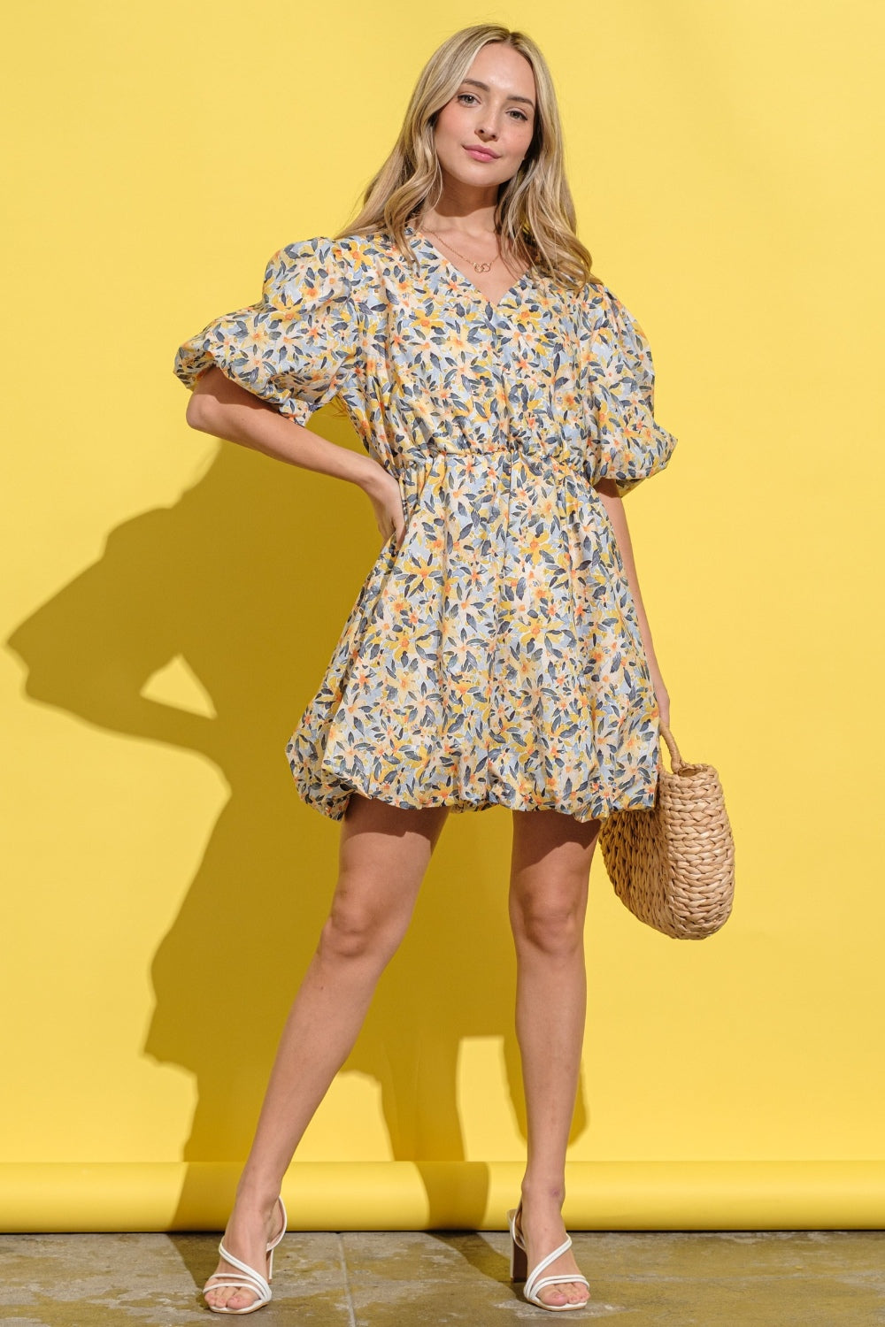 A person wearing the And The Why Full Size Floral Surplice Puff Sleeve Dress stands against a yellow background, holding a woven bag and wearing white sandals.