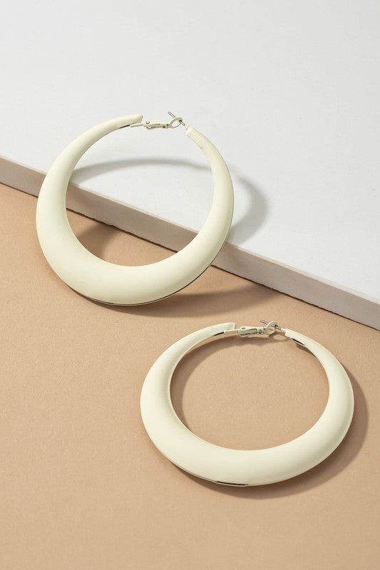 Four large color coated puffy hoop earrings, available in off-white, pink, blue, and black hues, are neatly displayed on a beige surface and white platform.