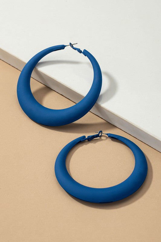 Four large color coated puffy hoop earrings, available in off-white, pink, blue, and black hues, are neatly displayed on a beige surface and white platform.