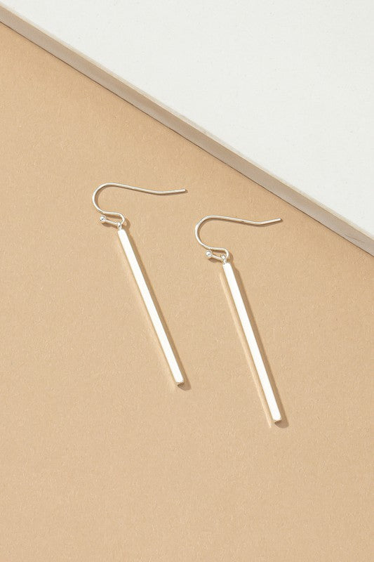 The "Minimalist Match Stick Drop Earrings" are elegantly displayed on a beige and white surface, with one pair showcasing luxurious gold plating and the other pair gleaming in silver.