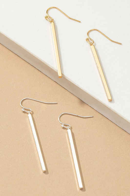 The "Minimalist Match Stick Drop Earrings" are elegantly displayed on a beige and white surface, with one pair showcasing luxurious gold plating and the other pair gleaming in silver.