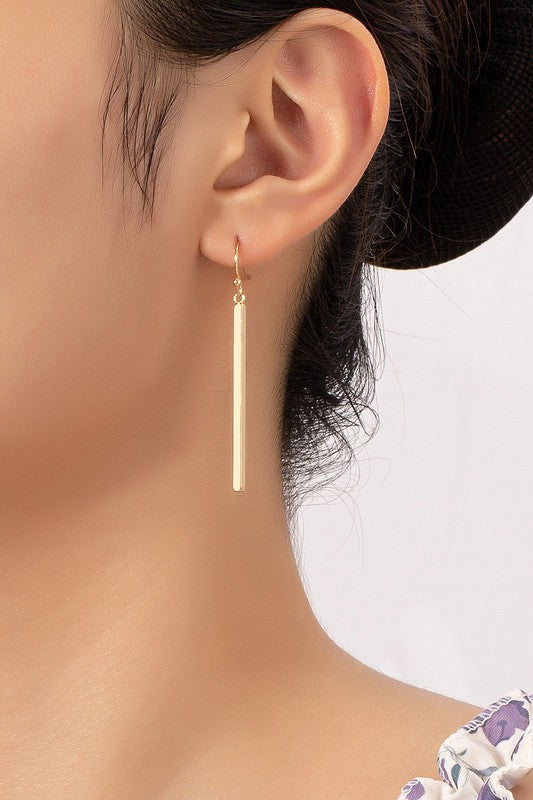 The "Minimalist Match Stick Drop Earrings" are elegantly displayed on a beige and white surface, with one pair showcasing luxurious gold plating and the other pair gleaming in silver.