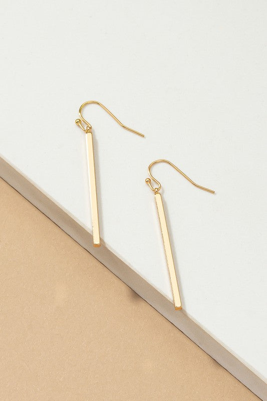 The "Minimalist Match Stick Drop Earrings" are elegantly displayed on a beige and white surface, with one pair showcasing luxurious gold plating and the other pair gleaming in silver.