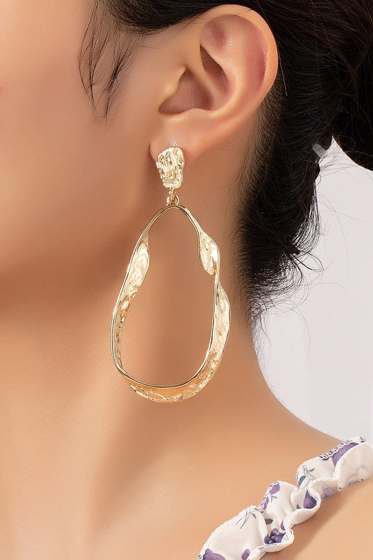 The Statement hammered twisted metal hoop earrings, with their asymmetrical shape, textured surfaces, and shiny gold plating, are elegantly displayed on a beige and white background, capturing the artisanal charm of their design.