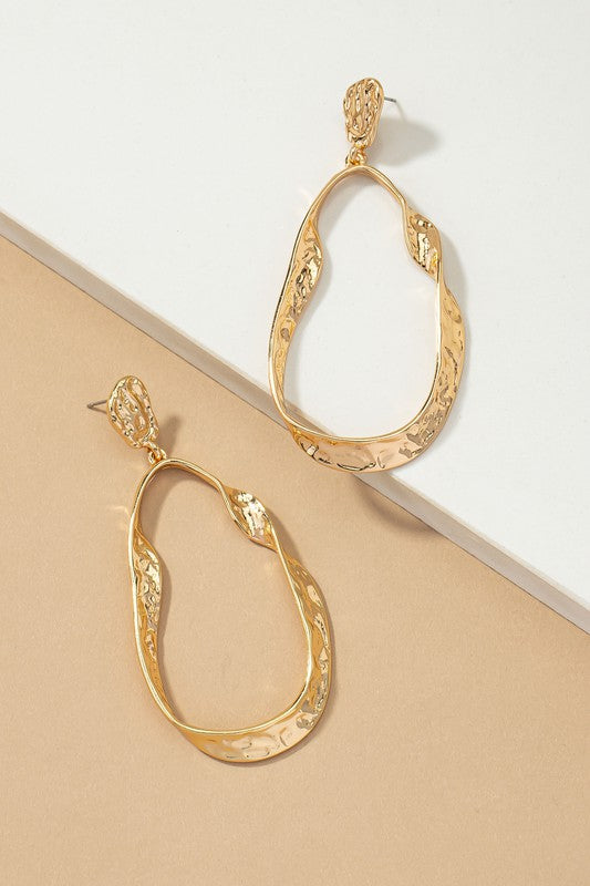 The Statement hammered twisted metal hoop earrings, with their asymmetrical shape, textured surfaces, and shiny gold plating, are elegantly displayed on a beige and white background, capturing the artisanal charm of their design.