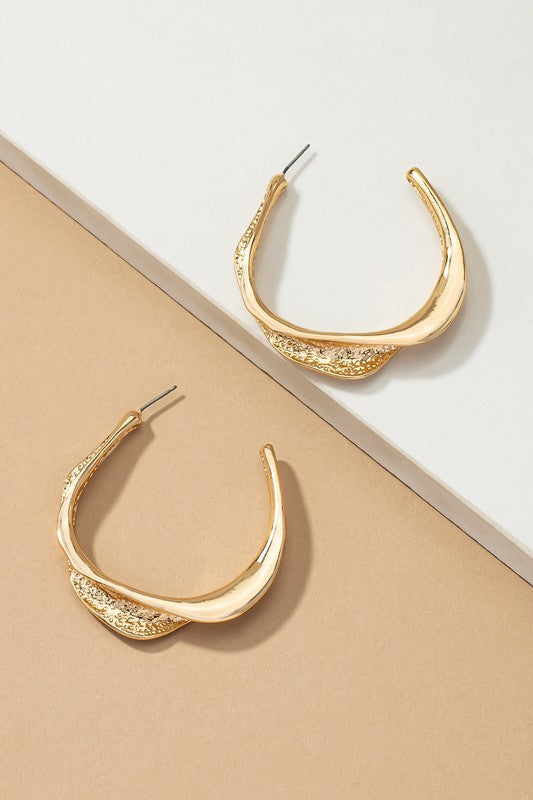 The Twisted Metal Hoop Earrings, with their gold hue and elegant crescent shape adorned with intricate twisted details, rest gracefully on a white and beige surface.