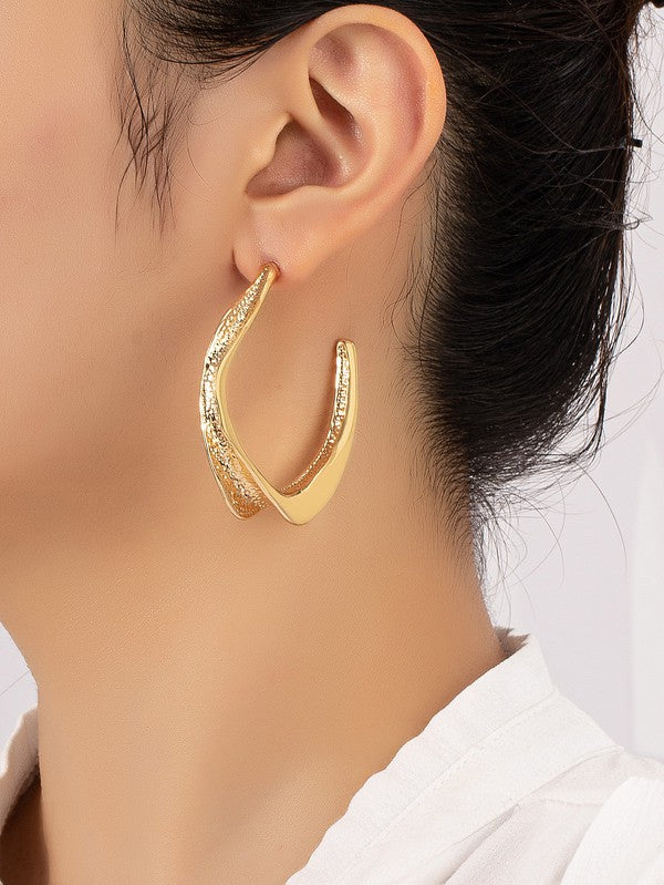 The Twisted Metal Hoop Earrings, with their gold hue and elegant crescent shape adorned with intricate twisted details, rest gracefully on a white and beige surface.