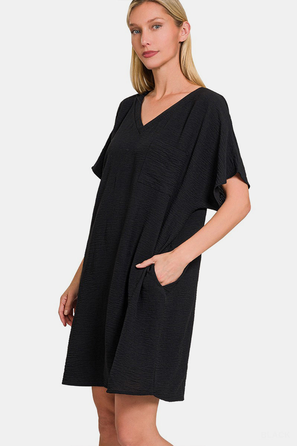 A woman with long hair is sporting the Zenana V-Neck Tee Dress with Pockets, a comfortable, loose-fitting black dress featuring short sleeves. She is smiling and has one hand on her hip.