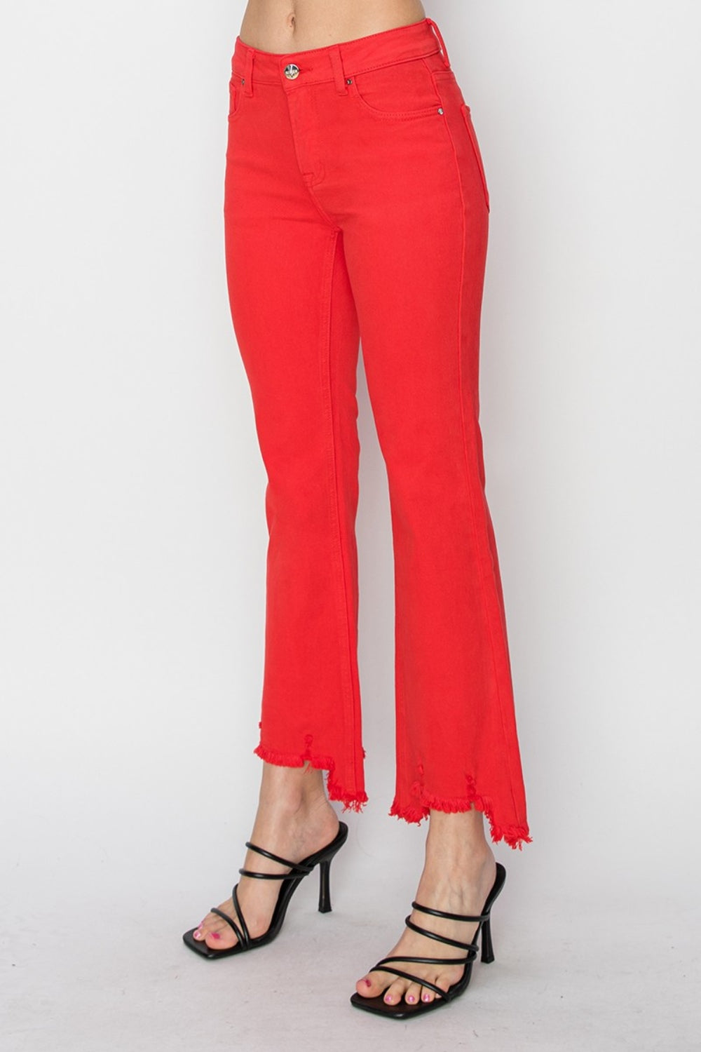 A person is wearing the RISEN Raw Hem Bootcut Jeans with Pockets in bright red, paired with a white top and black strappy high-heeled sandals. The image focuses on the stylish and functional lower half of the body.