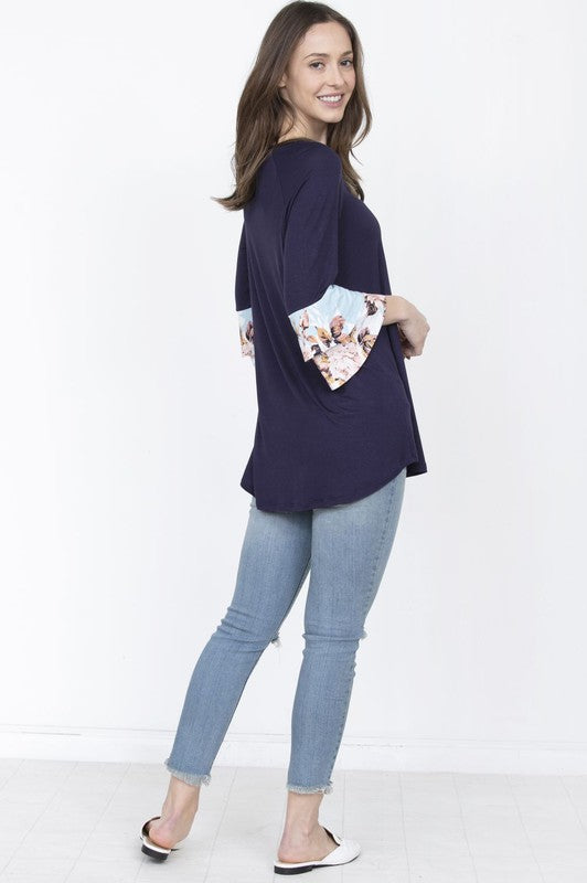 A woman wearing a Floral Double Ruffle Tunic with yellow fabric and floral-patterned sleeve cuffs stands against a beige curtain. Crafted with care in the United States, her style embodies a perfect blend of elegance and comfort.