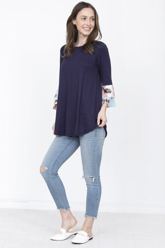 A woman wearing a Plus Floral Double Ruffle Tunic, featuring blue floral-patterned sleeves, stands in front of a beige curtain.